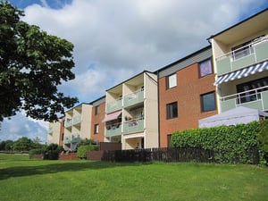 Lägenhetsbyggnad med röda tegelväggar och balkonger, omgiven av grönska och ett staket på en solig dag