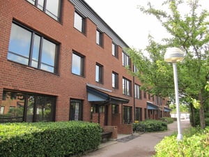 Röd tegelbyggnad med flera stora fönster och blåa entrétak, omgiven av grönska och en gångstig