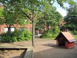 Lekplats med sand, ett rött lekhus och träbänkar, omgiven av grönskande träd och buskar framför röda tegelbyggnader