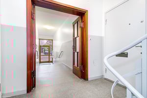 Entréområde med vita väggar, en trappa och en dörr i trä som leder till ytterligare en dörr med glaspaneler som öppnar mot utsidan