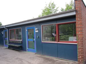 En enkel byggnad med blå fasad, röda fönsterkarmar och en blå dörr. Omgivningen har gröna träd och buskar