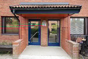 Entré till en tegelbyggnad med ett tak av röda tegelpannor, två glasdörrar, och en skylt med texten "22 C