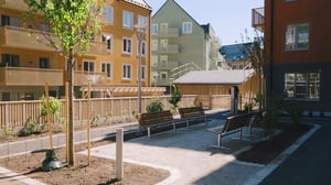 Modern bostadsområde med flerfamiljshus, parkbänkar, nyplanterade träd och ett staket