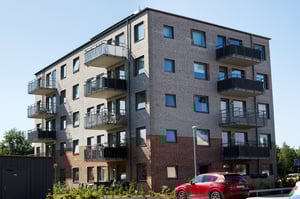 Flerbostadshus i tegel med balkonger, omgiven av grönska och en röd bil parkerad framför