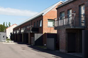 Radhus med fasad av rött tegel, balkonger i metall och svarta grindar, beläget vid en asfalterad väg under klarblå himmel