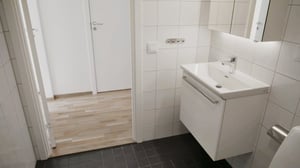 Ljust badrum med vita kakelväggar, vit kommod med spegel och svart klinkergolv, samt en öppning mot ett rum med parkettgolv