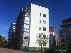 Modernt flerbostadshus med vita och röda fasader under en klarblå himmel, omgiven av träd och gångväg