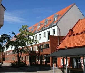 Byggnad med tegeltak och vita fasader under en klarblå himmel, omgiven av träd och en gård