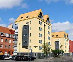 Gula flerfamiljshus med spetsiga tak i stadsmiljö, parkerade bilar och blå himmel