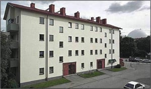 Flerfamiljshus med ljus fasad, röda dörrar och röda takpannor, beläget vid en gata med trottoarer och några parkerade cyklar