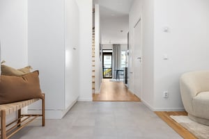 Hall med ljusa vita väggar, klinkergolv, en träbänk med kuddar till vänster och en beige fåtölj till höger. Utsikt mot en balkong genom glasdörrar i bakgrunden