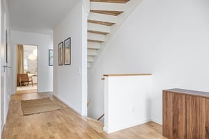 Ljust och modernt inrett hem med trägolv, vitmålade väggar, en trappa i trä och ett mörkt träskåp. Ett rum med matbord syns i bakgrunden