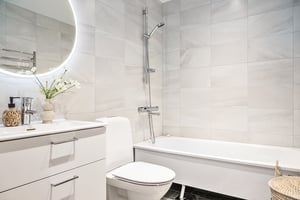 Modernt badrum med vita kakelväggar, rund spegel med belysning, toalett, badkar och handfat med dekorativ vas och tvålpump