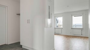 Interiör av en ljus lägenhet med vita väggar, ett garderobsskåp och parkettgolv. Fönster och radiatorer synliga längs väggen