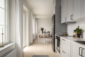 Modernt kök med vita skåp och rostfria vitvaror, anslutet till en matplats med ett dukat bord och stolar. Ljusa trägolv och stora fönster som släpper in naturligt ljus