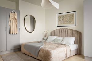 Sovrum med en beige säng, kudde och filt, en spegel på väggen, en inramad karta, och en beige klänning som hänger på en grå garderob