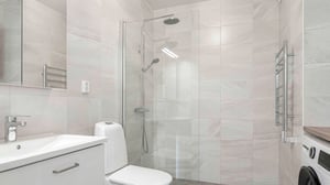 Modernt badrum med dusch bakom glasvägg, vit tvättställskommod, toalett, handdukstork och tvättmaskin. Väggar klädda i ljusa kakelplattor