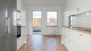 Ett modernt kök med vita skåpluckor, träbänkskivor och rostfria vitvaror, med utsikt mot en terrass genom två fönster