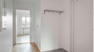Hall med vitmålade väggar, grått klinkergolv och en klädhängare. Öppen dörr leder till ett ljust rum med parkettgolv och fönster