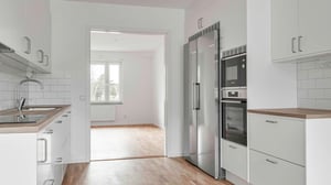 Modern kök med vita skåp, rostfria vitvaror och träbänkskivor, angränsande till ett rum med parkettgolv och stort fönster