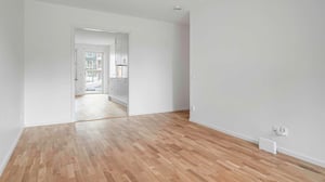 Inflyttningsklart rum med ljust trägolv och vita väggar, öppen planlösning mot ett kök i bakgrunden