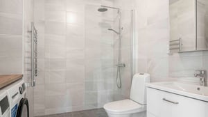 Badrum med duschhörna, toalett, handfat med lådor och tvättmaskin. Väggarna är kaklade och golvet har mörka plattor