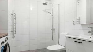 Badrum med vitkaklade väggar, dusch med glasvägg, toalett, tvättmaskin och vit handfat med spegelskåp