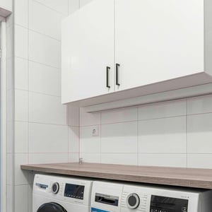 Tvättstuga med vitkaklade väggar, vit skåpförvaring, och två moderna tvättmaskiner på en träbänkskiva