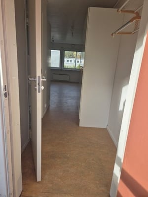 Vy in i ett rum med en öppen dörr, ljusa väggar, trägolv och ett fönster med utsikt