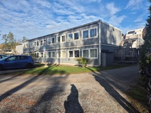 Grå byggnad i två våningar med flera fönster, omgiven av grusväg och parkerade bilar under en blå himmel