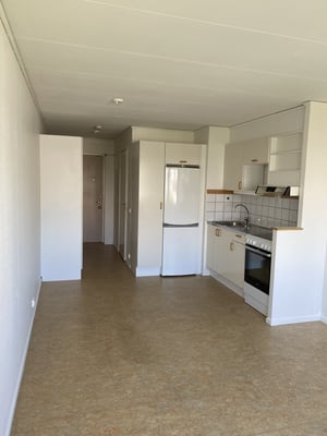 Litet kök med vit kyl, spis, ugn och diskho, intill en tom hall med vita väggar och trädörr