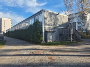 Grå byggnad med träpanel, omgiven av grusväg och grönska, med trappor på sidan. Bakom byggnaden syns fler bostadshus och träd i höstlandskap