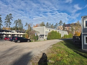 Parkeringsplats med bilar framför en låg byggnad, omgiven av träd och flerbostadshus under en blå himmel med moln
