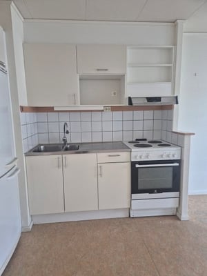 Litet kök med vita skåp, diskho, spis och ugn. Vägg med kakel ovanför köksbänken