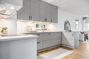 Modernt kök med gråa skåp, vit bänkskiva och kaklade väggar. Trägolv och en ljusmatta syns i förgrunden, med vardagsrum i bakgrunden