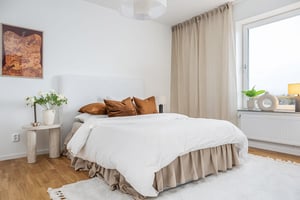 Sovrum med en stor säng klädd i vitt och beige sängkläder, vita väggar, ett fönster med beige gardin, och en träpanel på golvet. En nattduksbord med blommor och en tavla på väggen