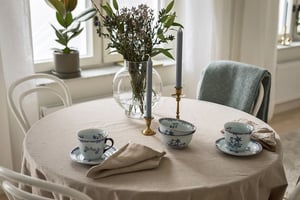 Rund matbord med duk, porslinskoppar med blått mönster, ljusstakar och en vas med blommor, omgiven av stolar i ett ljust rum