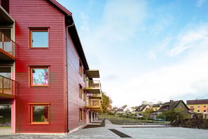 Flerfamiljshus med röd träfasad, balkonger och omgivande trädgårdar, med hus och blå himmel i bakgrunden