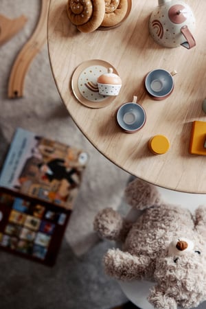 Ett runt träbord med porslinsservis i pastellfärger, små bröd, leksaksdelar och en mjuk teddybjörn på golvet bredvid ett pussel