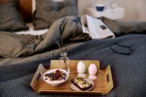 Frukostbricka med en skål yoghurt toppad med färska bär, två löskokta ägg, en smörgås och en liten vas med lavendel på ett träfat, placerad på en säng med grå sängkläder