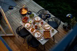 Middag på en träaltan med grill och matbord, omgiven av grönska. Bordet är dukat med tallrikar, drycker, grönsaker, bröd och tillbehör
