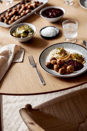 Tallrik med köttbullar, potatismos, lingonsylt och inlagd gurka på ett träbord, omgiven av andra skålar med mat och ett glas vatten