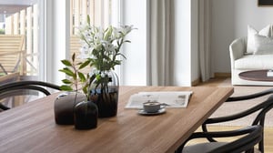 Blombukett i mörka vaser på ett träbord, med en kaffekopp och en bok, omgiven av moderna möbler och stora fönster med ljusinsläpp