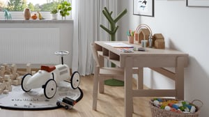 Ljust barnrum med träbord, leksaksbil, byggklossar och förvaring. Gröna växter och dekorationer syns vid fönstret