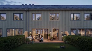 Radhus i trä med två våningar, omgivna av grönska och en trädgård; människor umgås på uteplatsen vid skymning