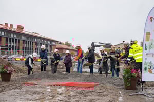 Personer klädda i byggarbetskläder står på en byggarbetsplats med spadar, framför en röd matta och en skylt. Bakgrunden visar en byggnad och en grävmaskin
