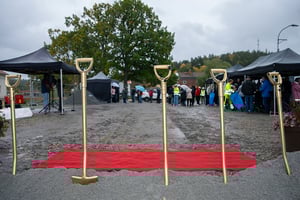 Fyra guldfärgade spadar på rad framför en röd matta på en byggarbetsplats, med tält och människor i bakgrunden under mulen himmel