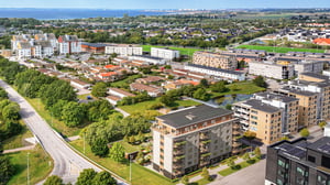 Flygvy över ett bostadsområde med flerfamiljshus, grönområden och vägar, med havet synligt i bakgrunden