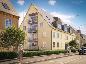 Gult flerfamiljshus med balkonger, omgiven av träd och en trottoar under en klarblå himmel