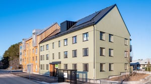 Flerbostadshus med modern design, grön fasad och solpaneler på taket, beläget vid en asfalterad gata med klarblå himmel